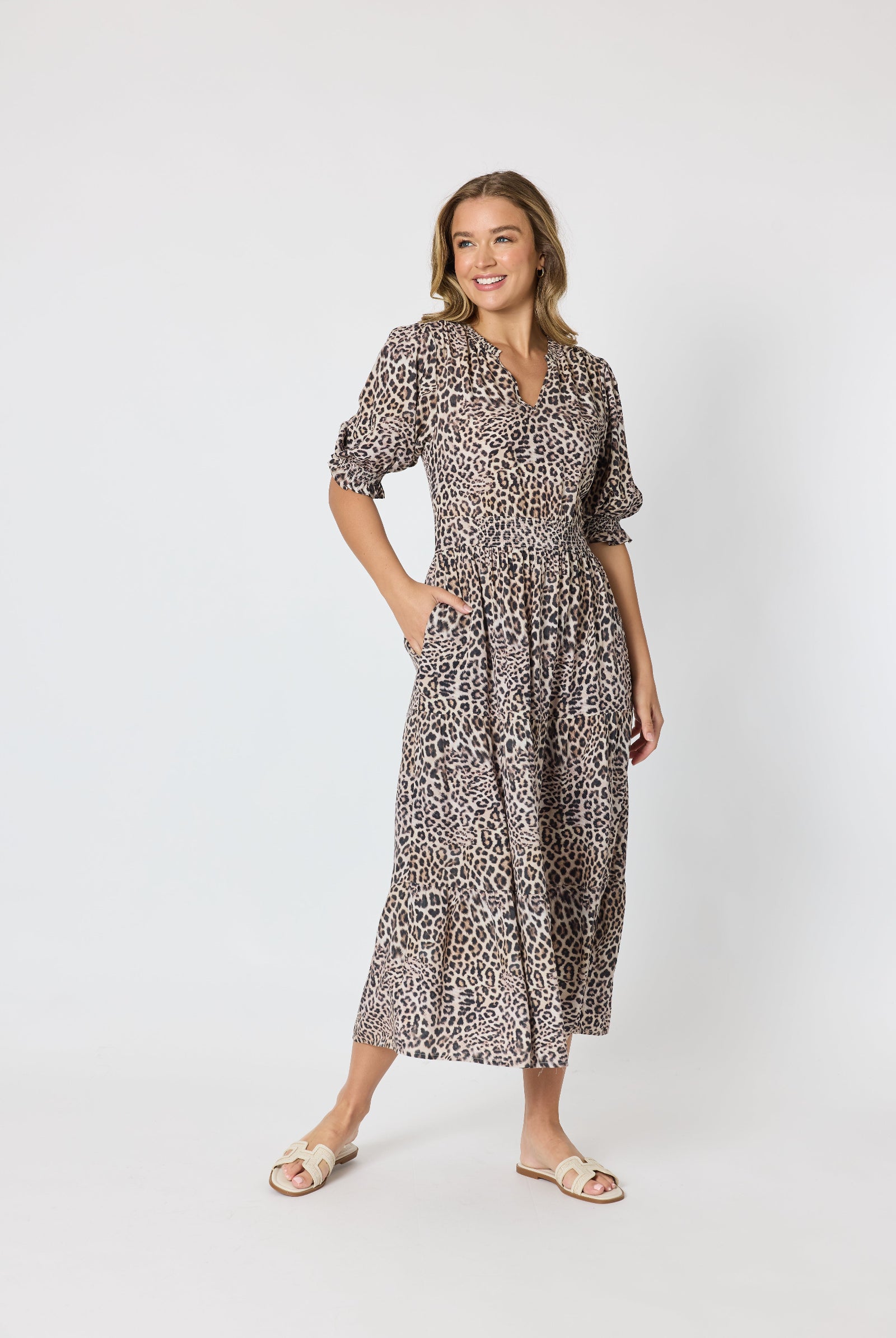 A woman modeling a leopard-print midi dress with puff sleeves and a V-neckline, standing against a plain light gray studio background.