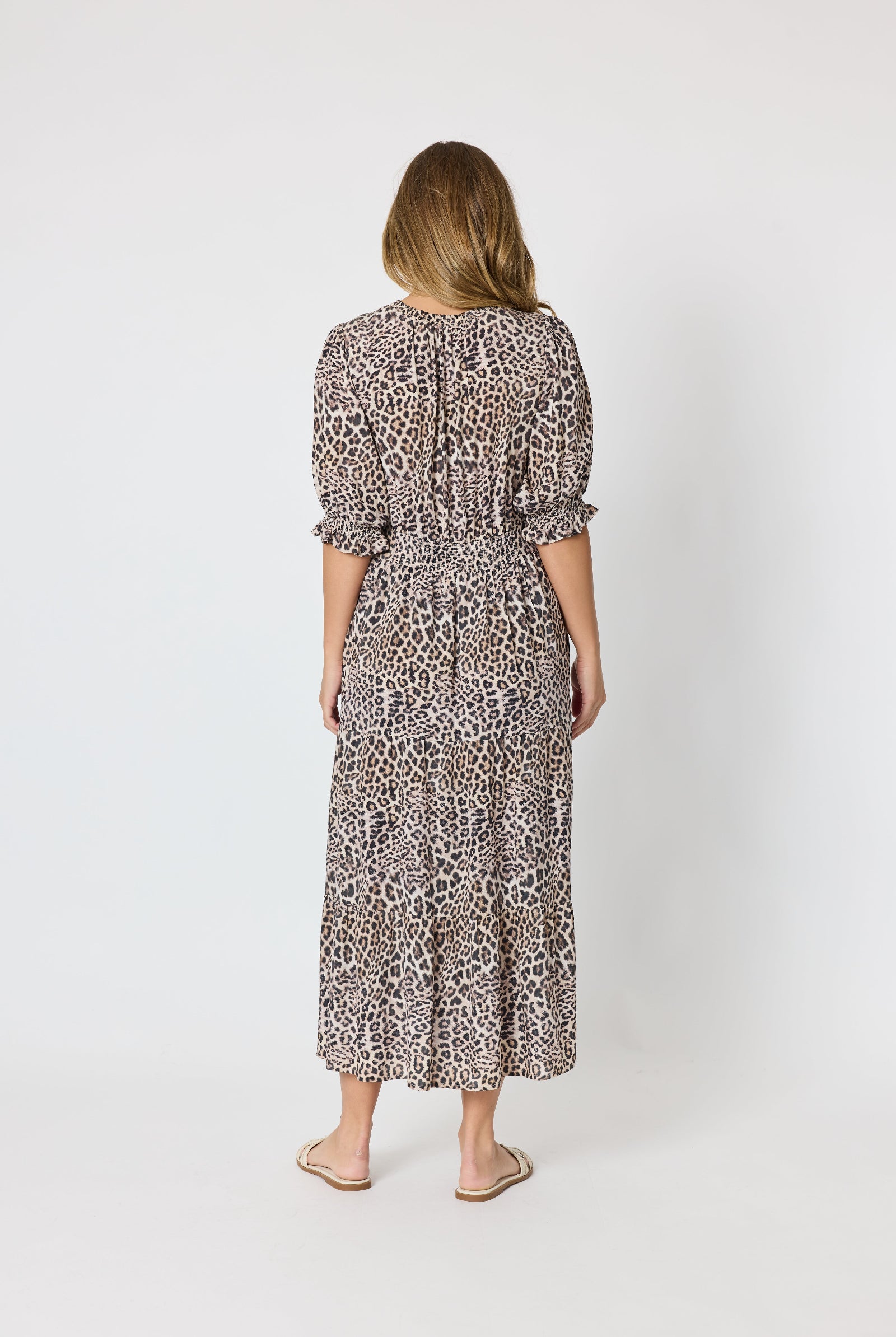 A woman viewed from behind wearing a leopard-print midi dress with puff sleeves and a tie at the back, paired with beige flat sandals, against a plain light gray studio background.