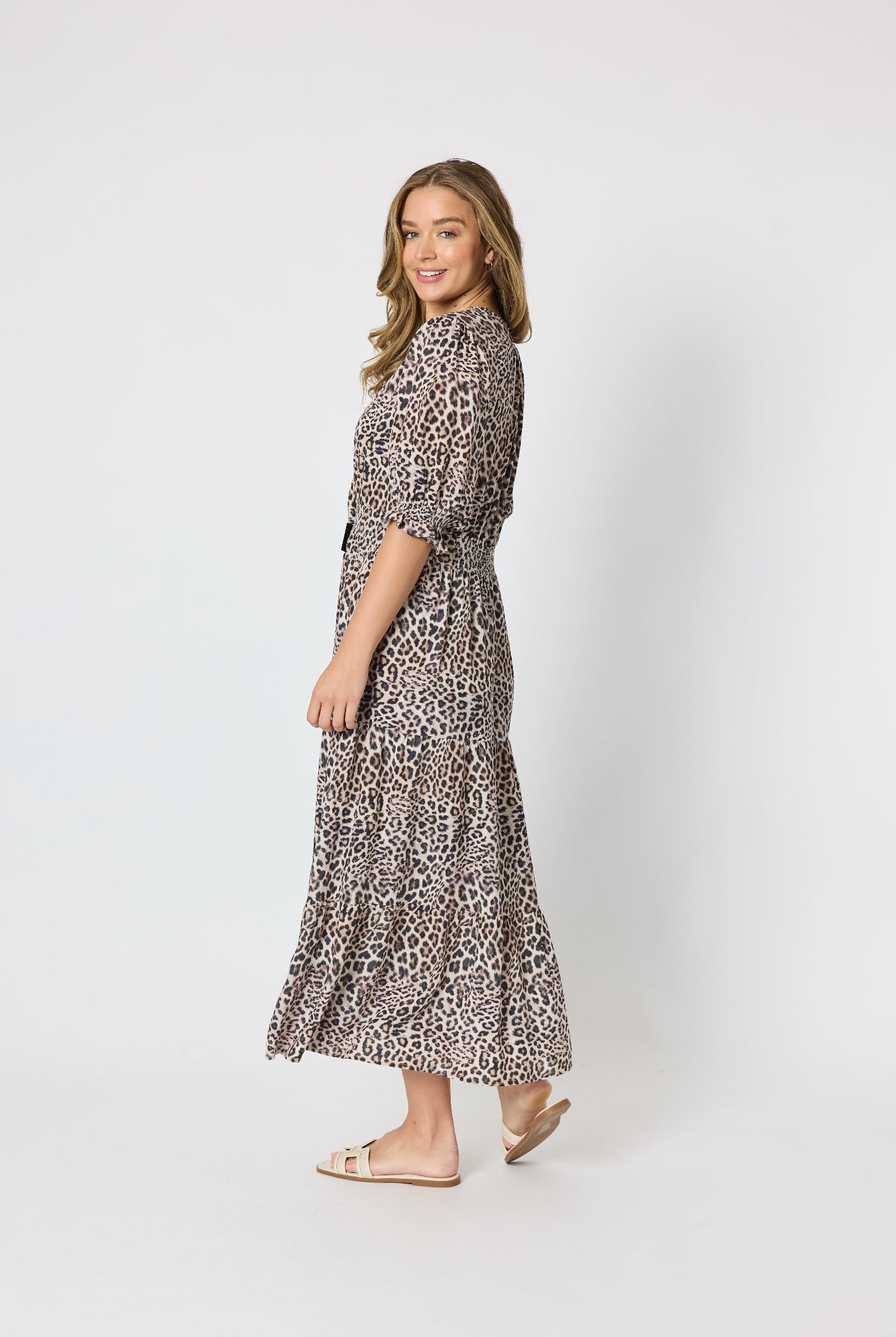 A woman modeling a leopard-print midi dress with three-quarter sleeves and a matching tie belt, standing against a plain light gray studio background.