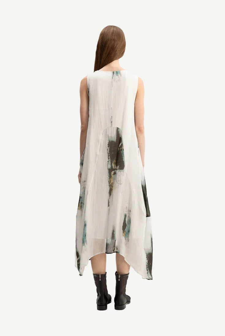 A woman viewed from behind wearing a sleeveless, high-low midi dress with an abstract watercolor print in green, black, and gray on a white base, paired with black ankle boots, against a solid off-white background.