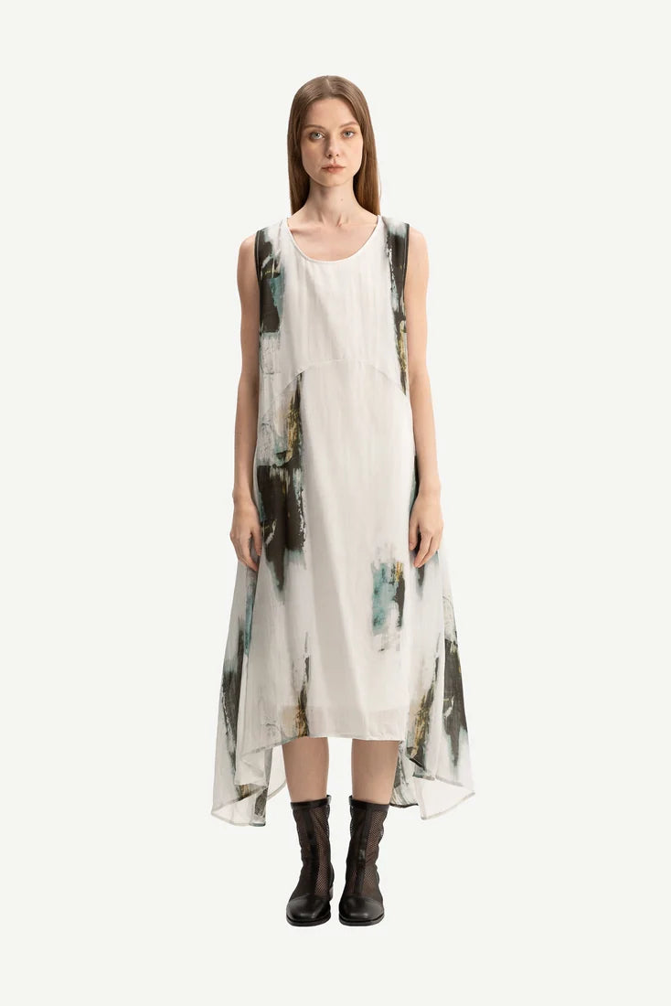 A woman modeling a sleeveless high-low midi dress with an abstract painterly print in black, gray, and teal on a white base, paired with black ankle boots, against a solid off-white background.