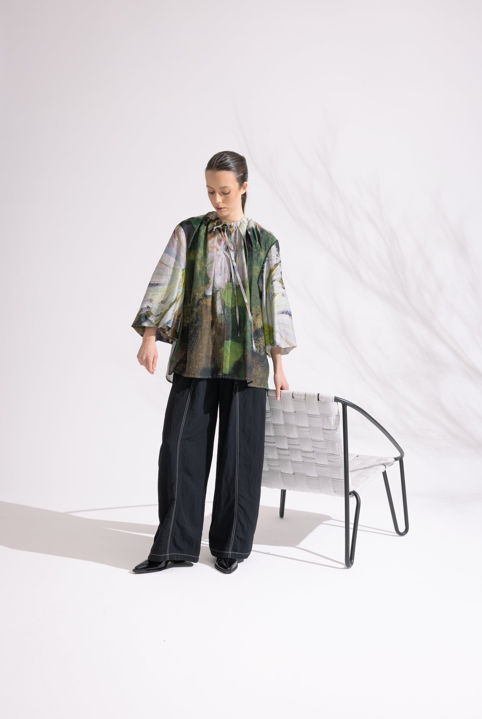Person wearing a patterned shirt and black pants standing in a minimalistic setting with a chair.
