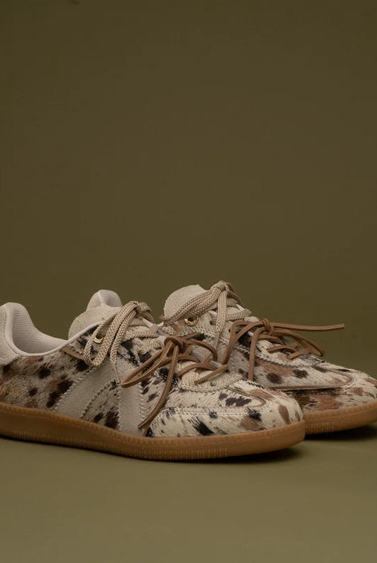 A pair of low-top sneakers with a beige, brown, and black spotted animal print upper, white laces, and gum soles, set against a solid olive green background.