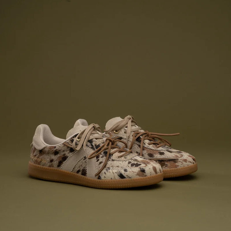 A pair of low-top sneakers with a beige, brown, and black spotted animal print upper, white laces, and gum soles, set against a solid olive green background.