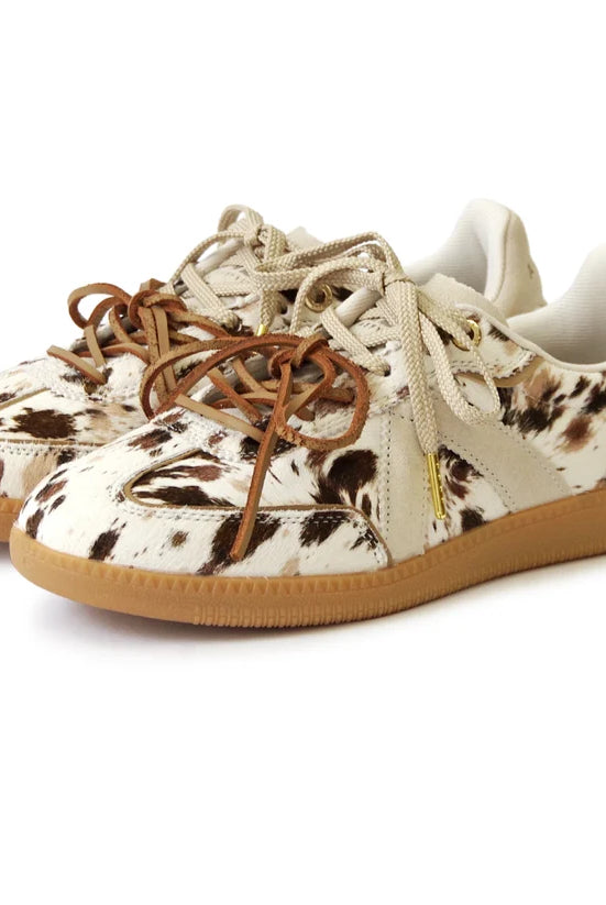 A pair of low-top sneakers featuring a cowhide-textured pattern in cream, brown, and black, with tan laces and gum soles, shown on a white background.