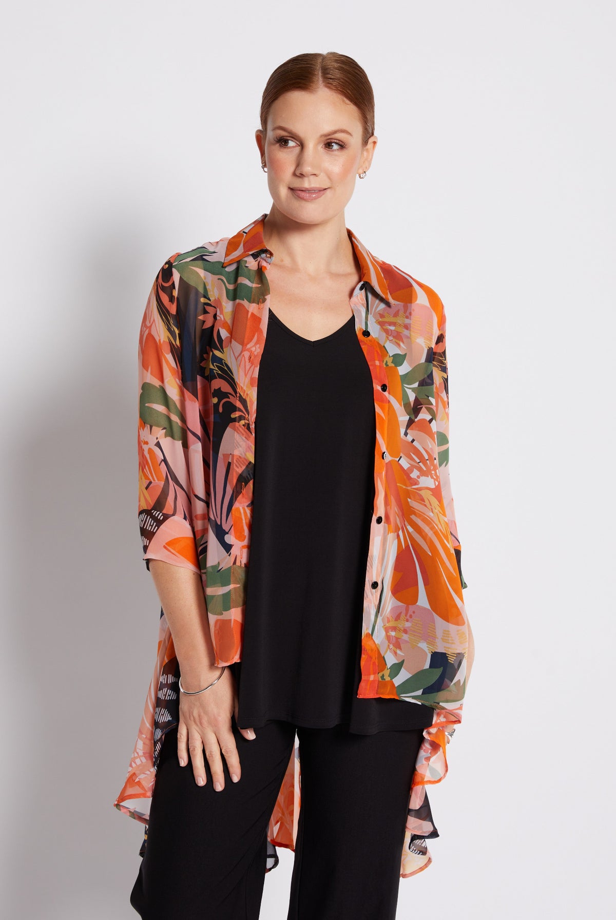A woman modeling a lightweight, semi-sheer button-up shirt with a vibrant orange, pink, and green abstract floral print over a black top and black pants, set against a plain light gray studio background.