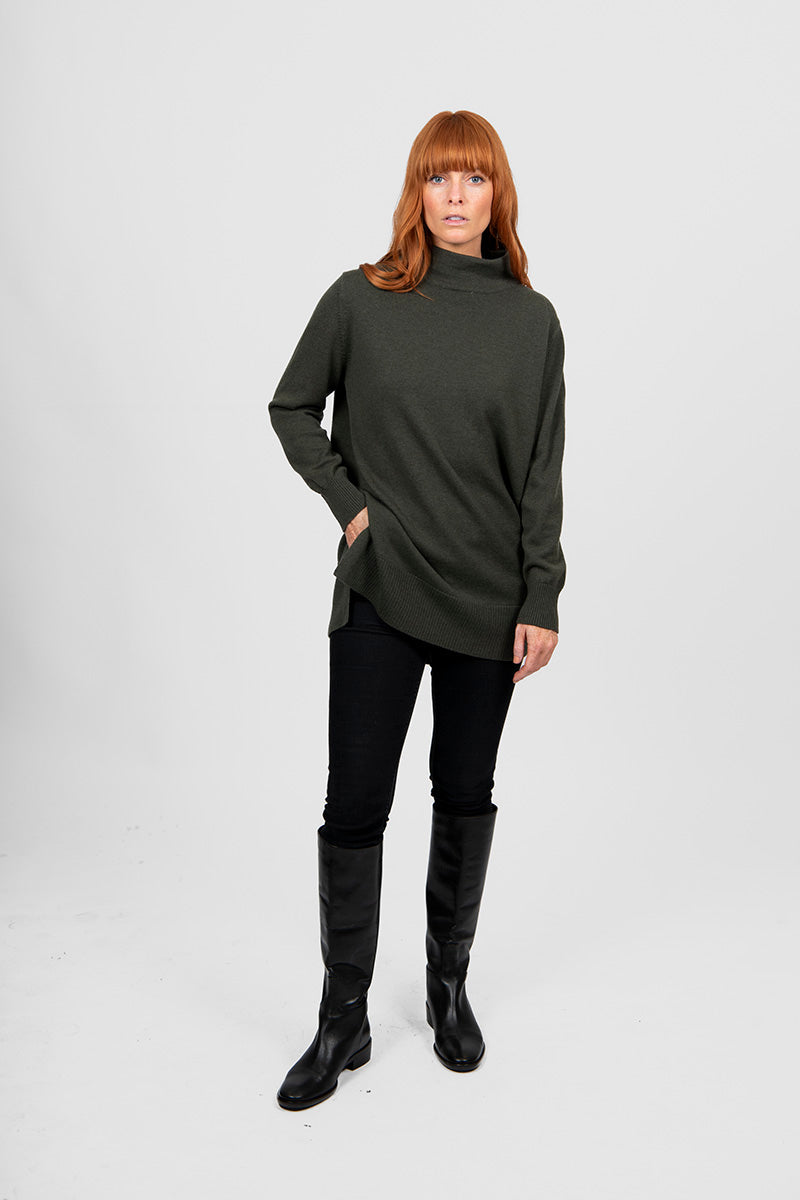 Woman wearing a long olive green sweater and black leggings with knee-high boots on a white background