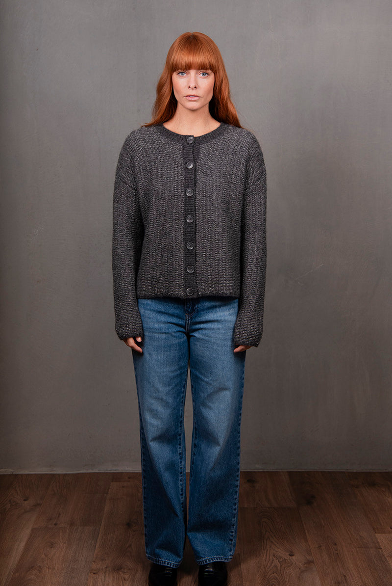 Woman wearing a gray textured cardigan, blue jeans, and black shoes against a gray background