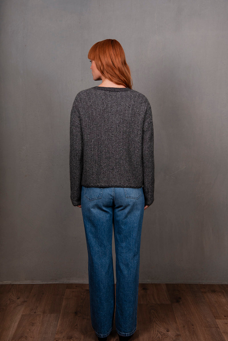 Person wearing a gray sweater and blue jeans standing against a plain gray wall.