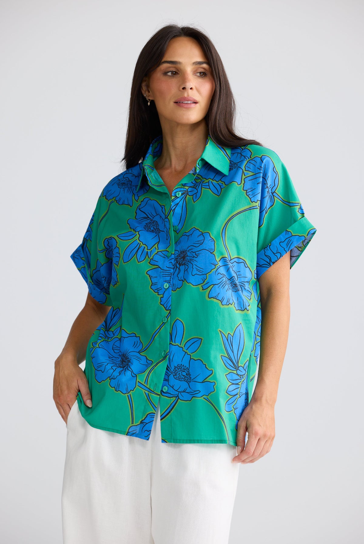 A woman modeling a short-sleeve button-up shirt with a vibrant green base and a bold blue floral pattern outlined in gold, paired with white pants against a light gray studio background.