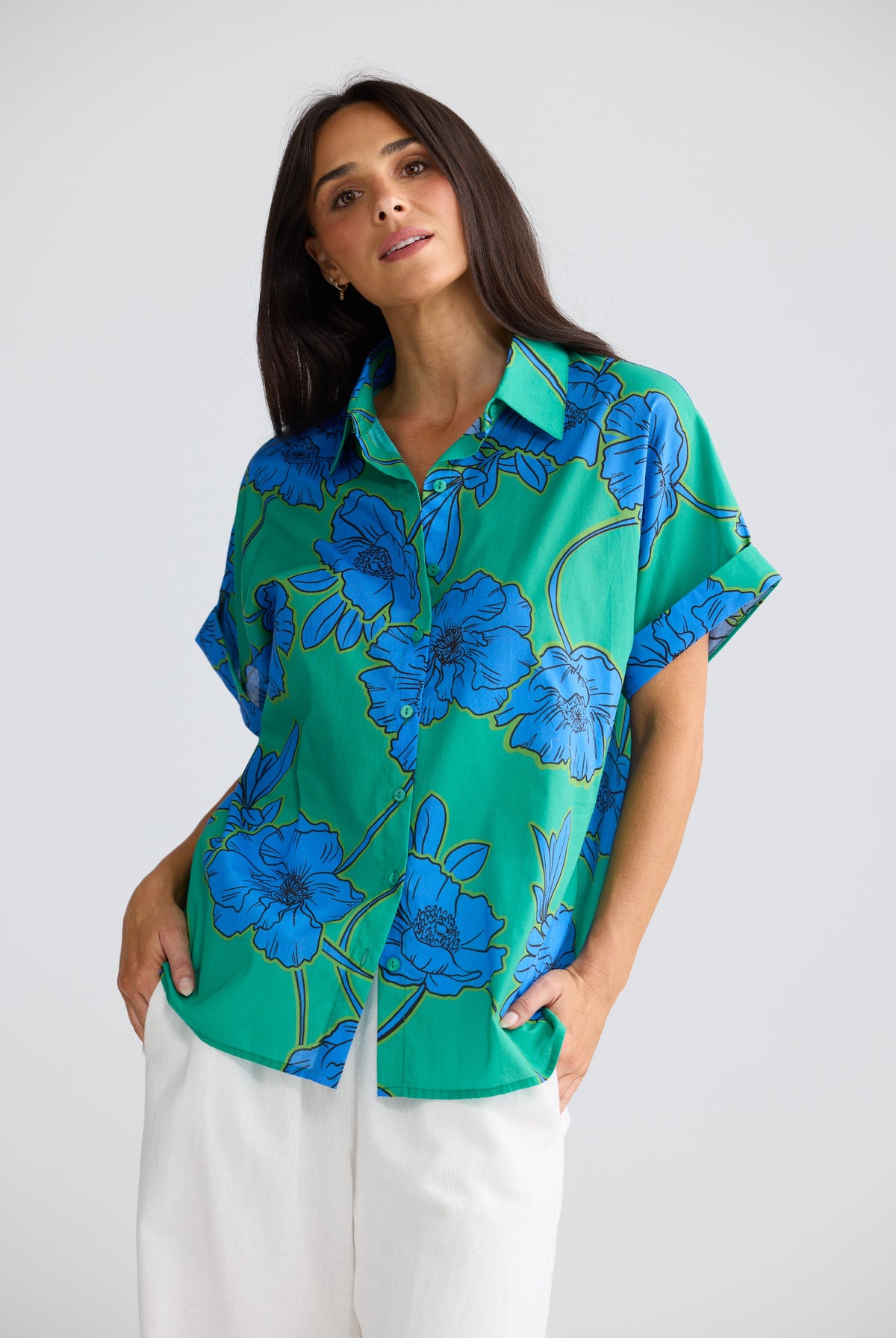 A woman wearing a vibrant green short-sleeve button-up shirt with a bold blue floral print, paired with white pants, photographed against a neutral light gray background.
