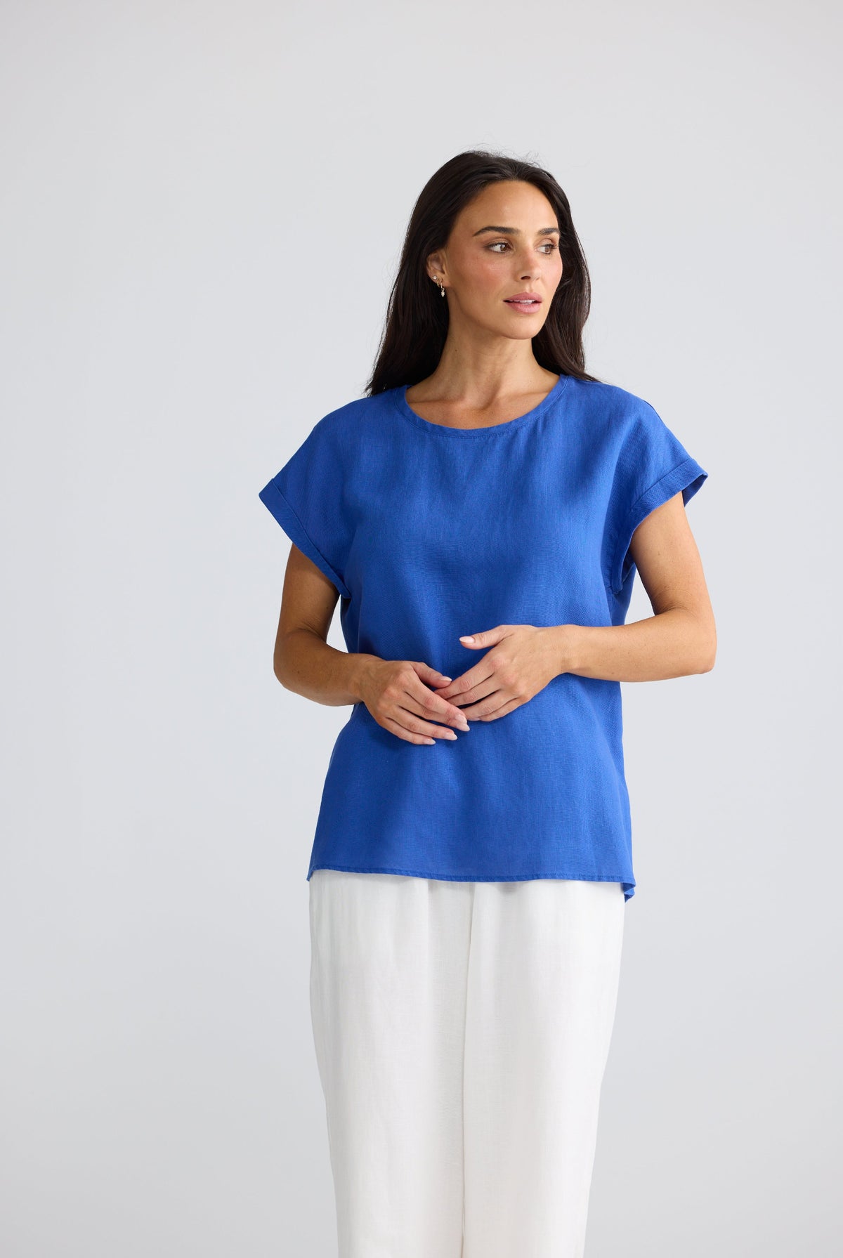 A woman modeling a bright blue short-sleeve top with white pants, standing against a light gray studio background.