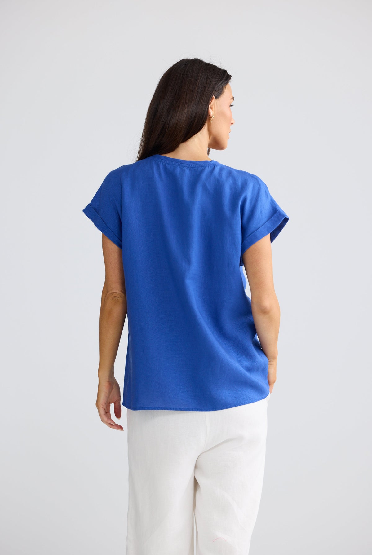 A woman shown from the back wearing a bright royal blue short-sleeve top and white pants against a light gray studio background.