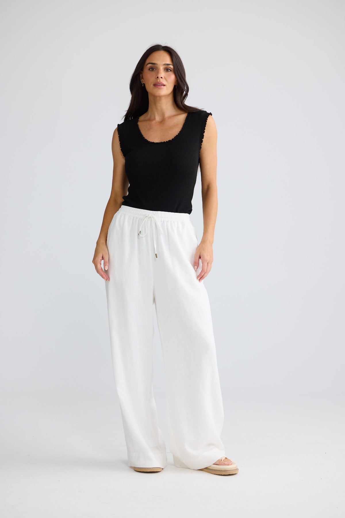 A woman modeling a black sleeveless scoop-neck top and white wide-leg pants against a light gray studio background.