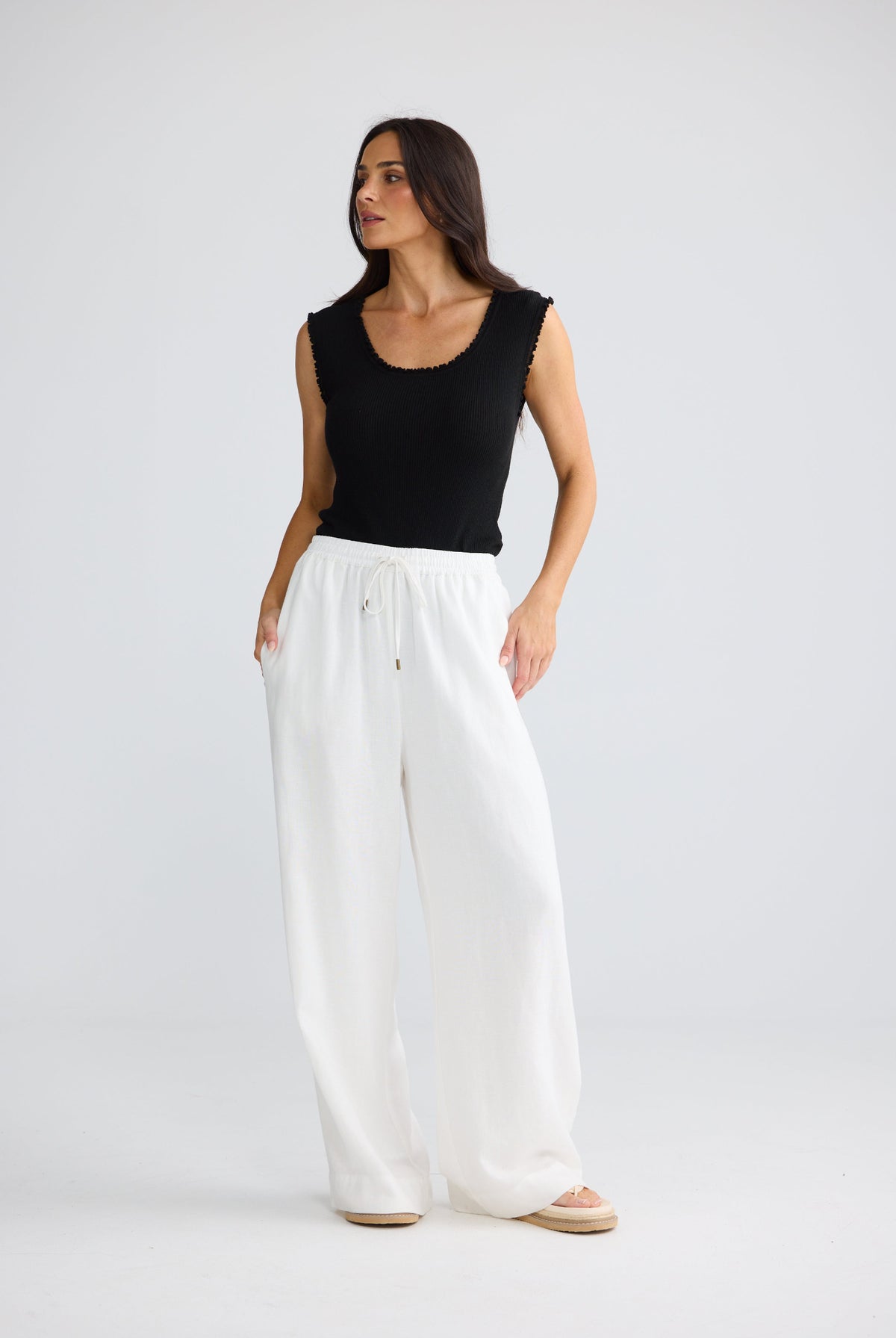 A woman modeling a black sleeveless scoop-neck top and white wide-leg drawstring pants against a light gray studio background.