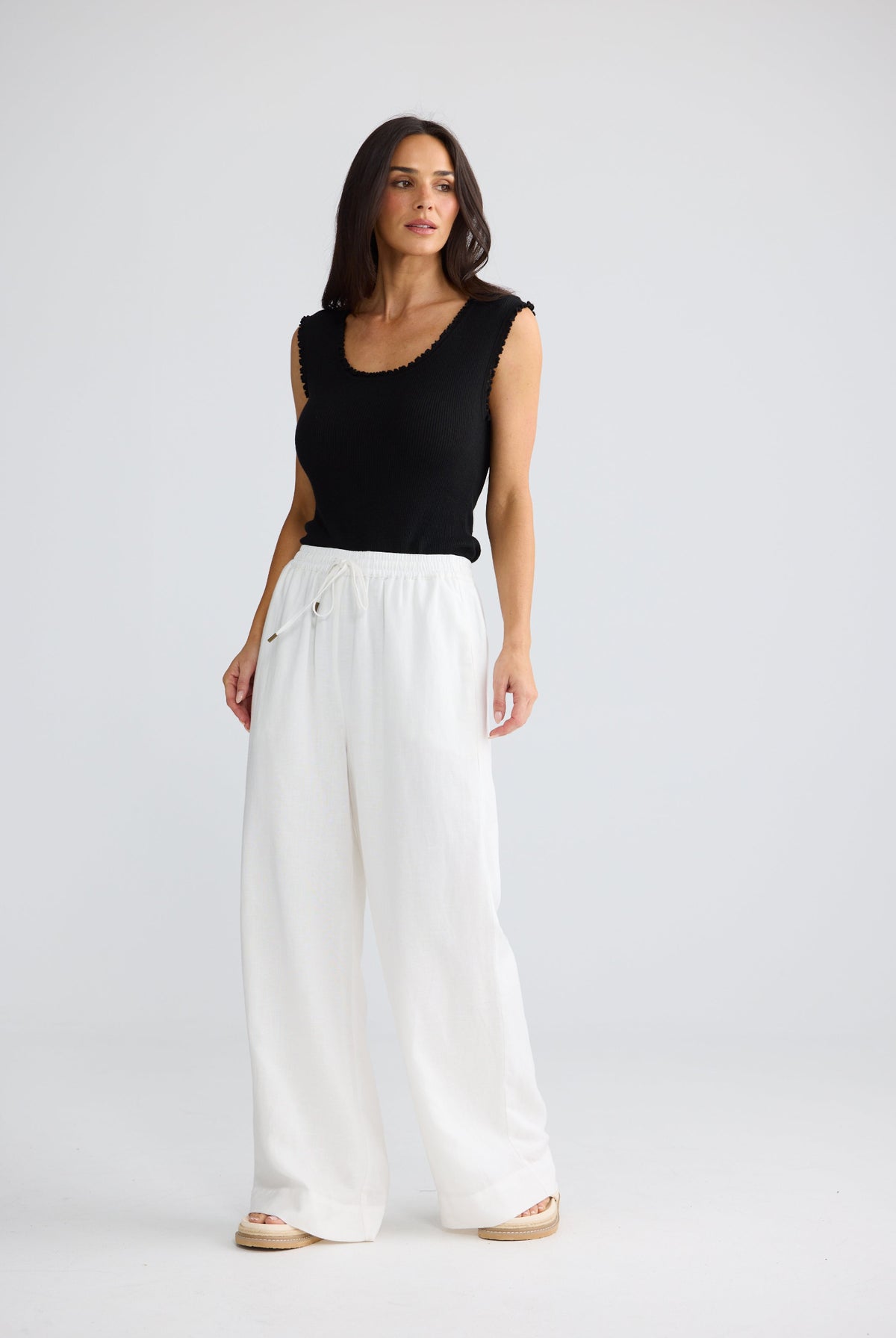 A woman modeling a black sleeveless top and white wide-leg pants against a light gray studio background.