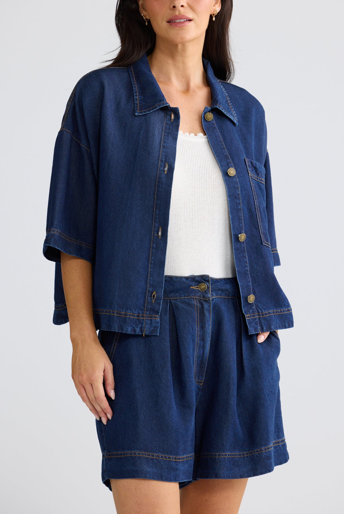 A woman wearing a dark blue denim jacket with three-quarter sleeves and matching denim shorts, styled over a white ribbed top, against a light gray studio background.