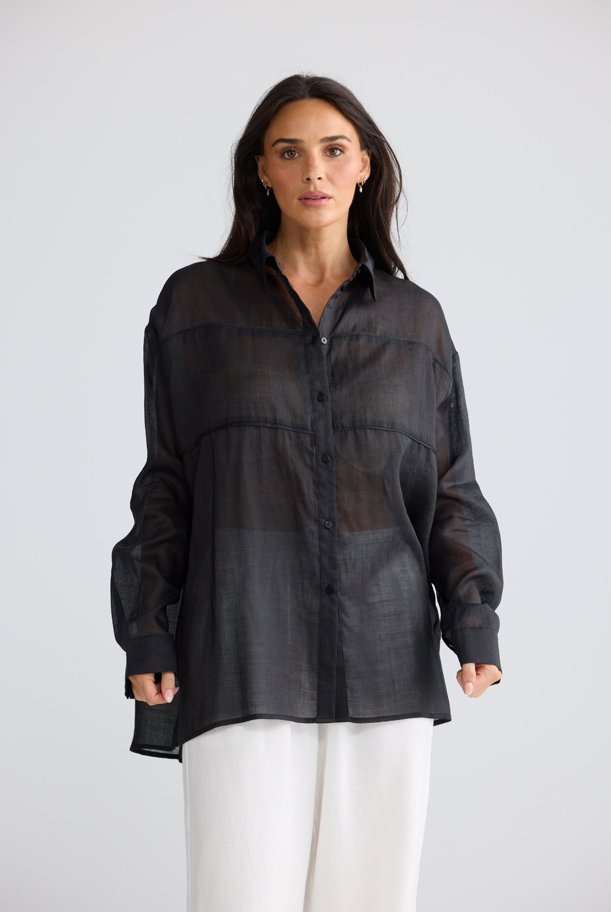 A woman modeling a sheer black long-sleeve button-up shirt with horizontal seam detailing, paired with white pants, against a light gray studio background.