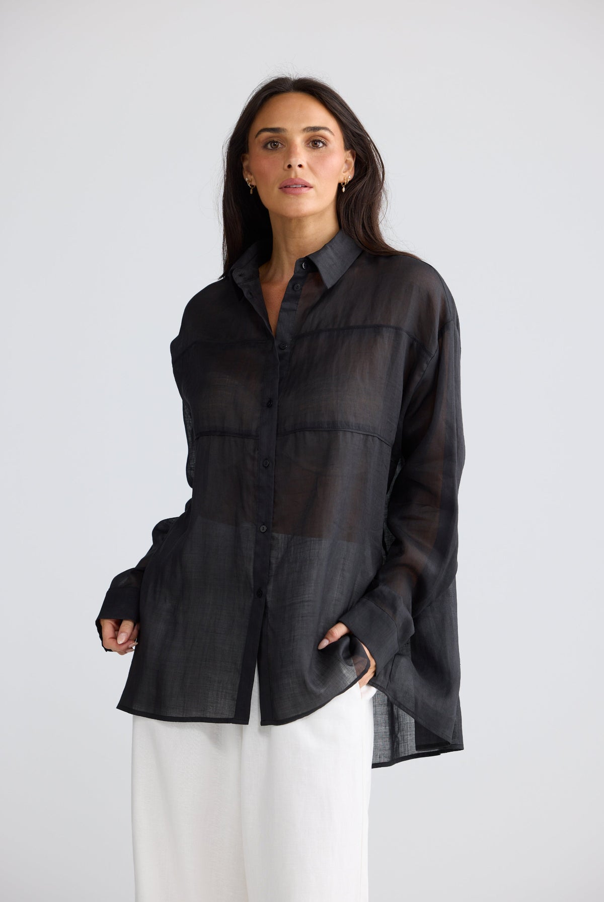 A woman modeling a sheer black long-sleeve button-up shirt with a relaxed, slightly oversized fit, paired with white pants, against a light gray studio background.