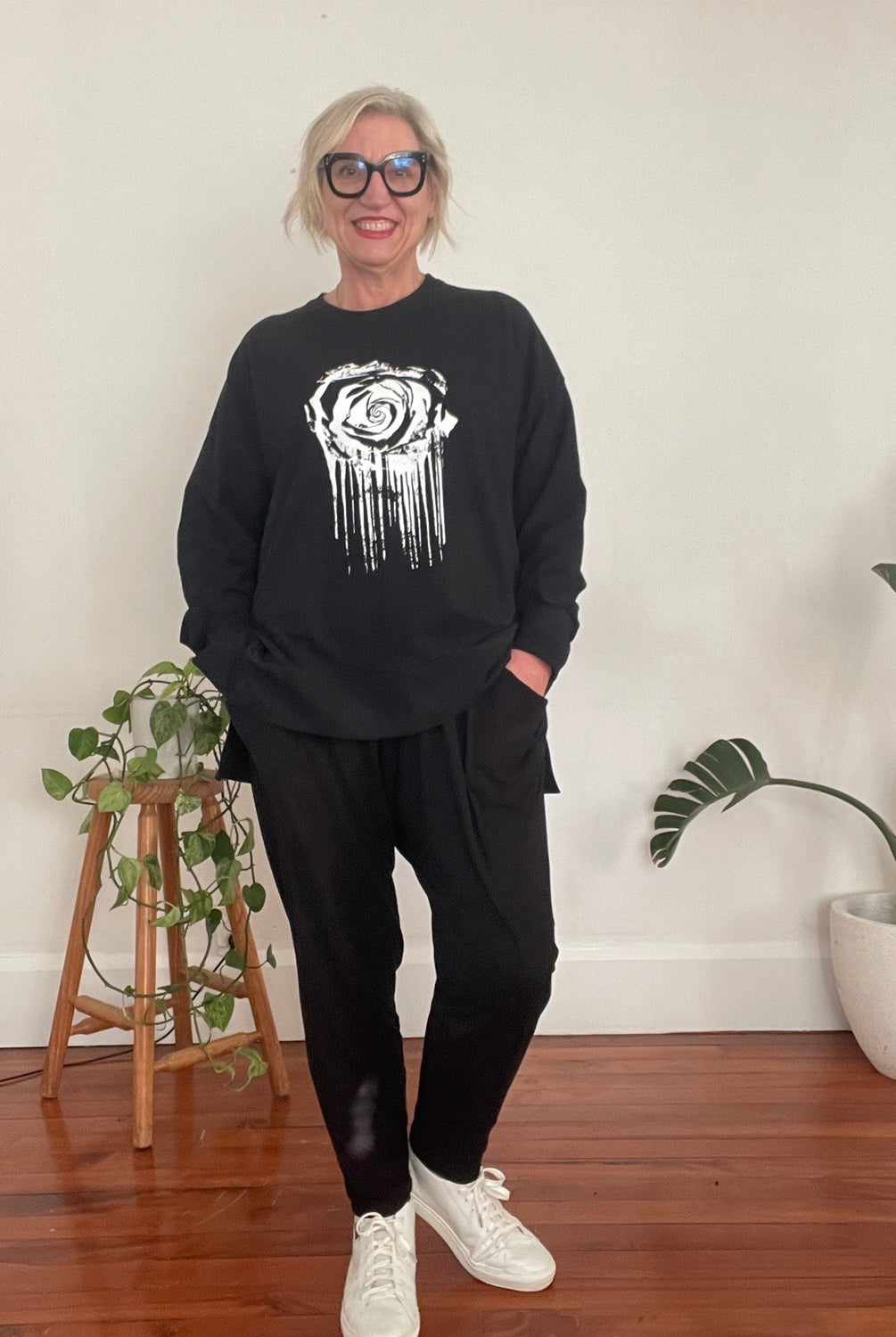 Person wearing a black sweatshirt with a white graphic design, black pants, and white sneakers in a room with plants and a vase.