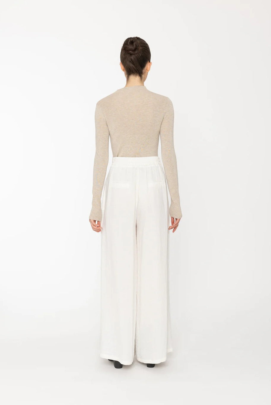 Person wearing a beige long-sleeve top and white wide-leg pants on a white background back angle