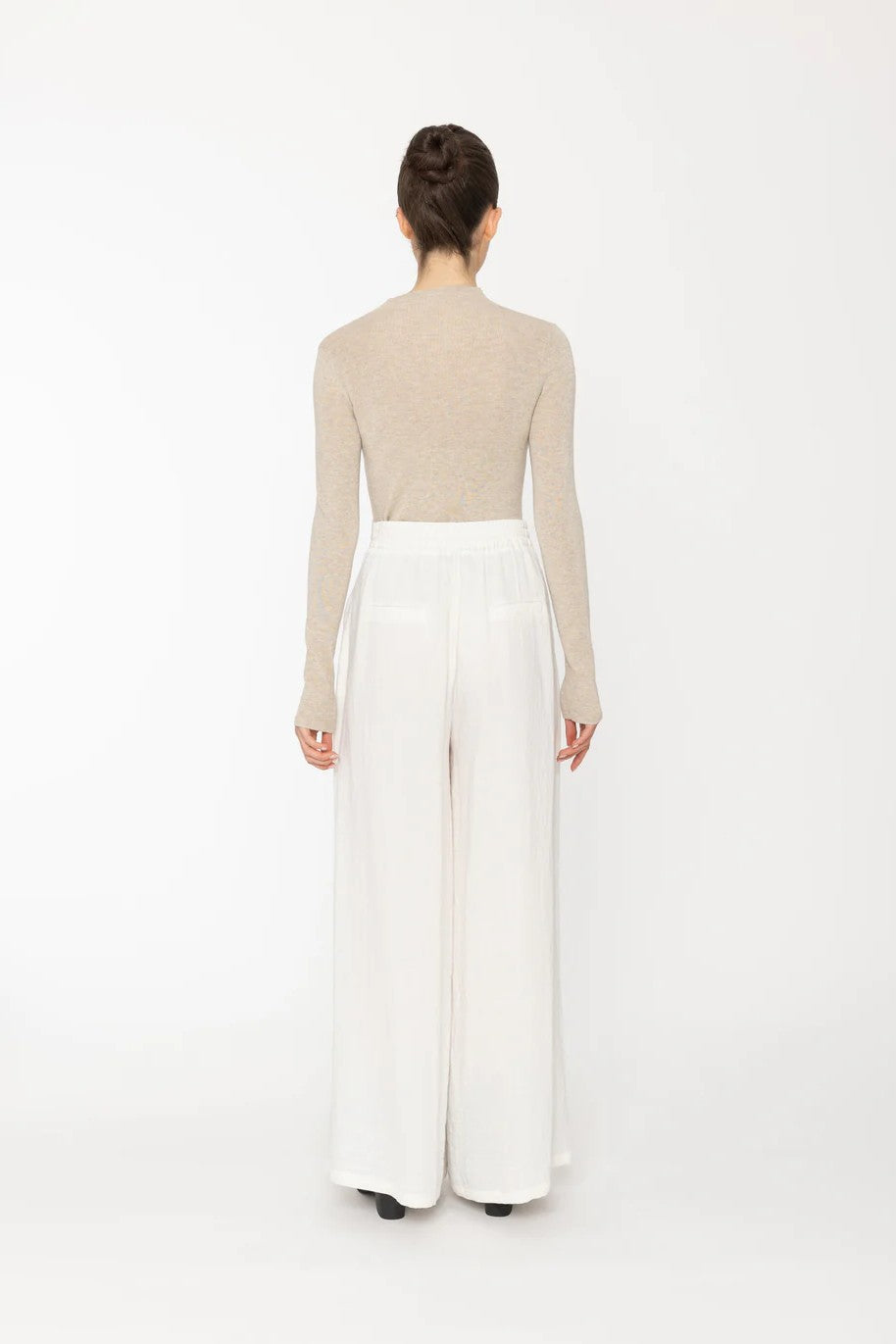 Person wearing a beige long-sleeve top and white wide-leg pants on a white background back angle