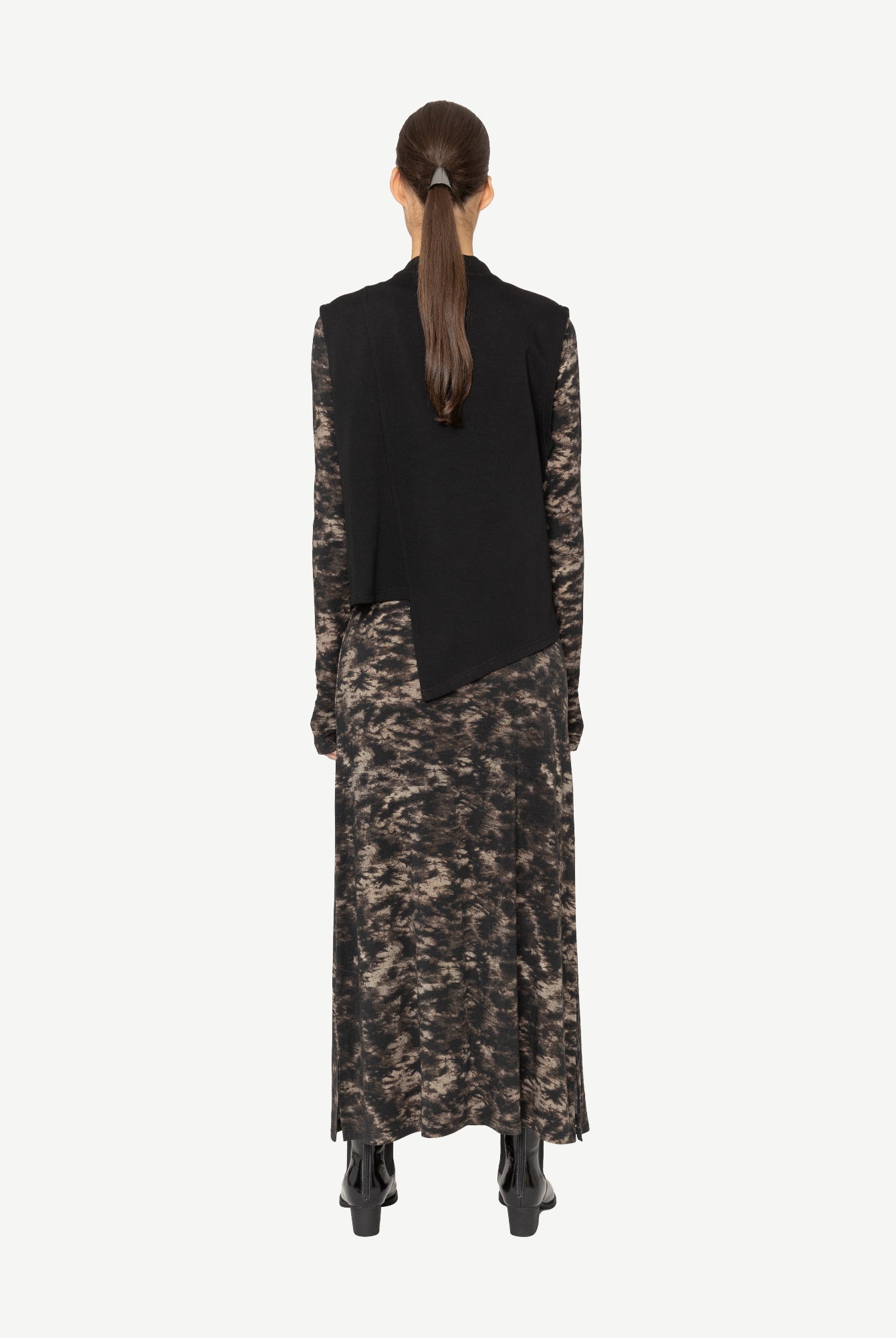 Person wearing a black vest over a patterned dress on a white background back angle