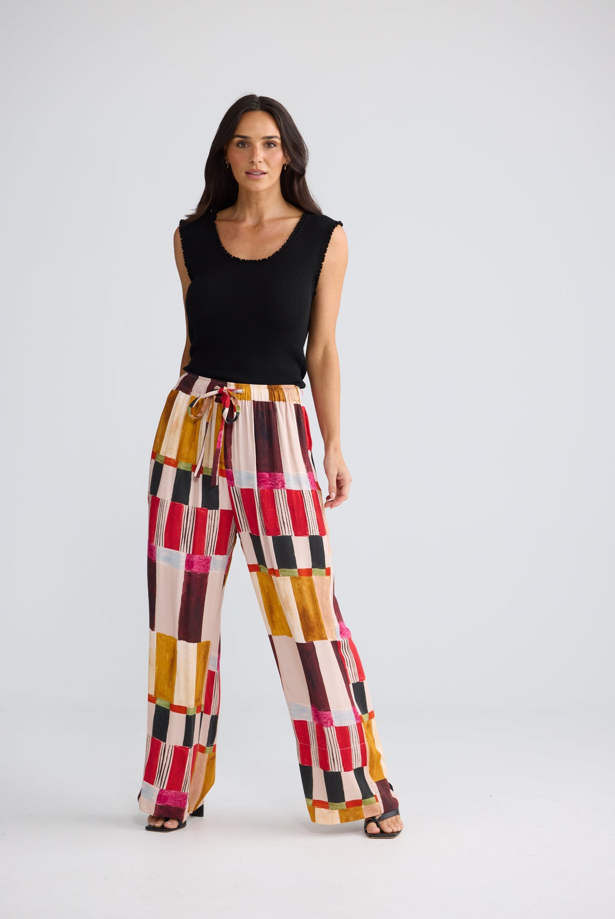 A woman modeling a pair of wide-leg pants with a colorful geometric patchwork pattern in pink, red, yellow, brown, and white, paired with a black sleeveless top, against a light gray studio background.