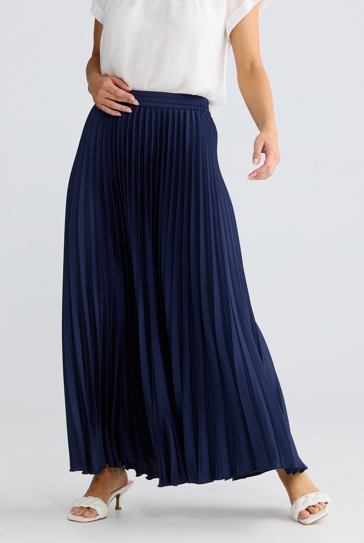 A navy blue pleated maxi skirt worn by a model, styled with a white sleeveless top and white heeled sandals against a light gray studio background.