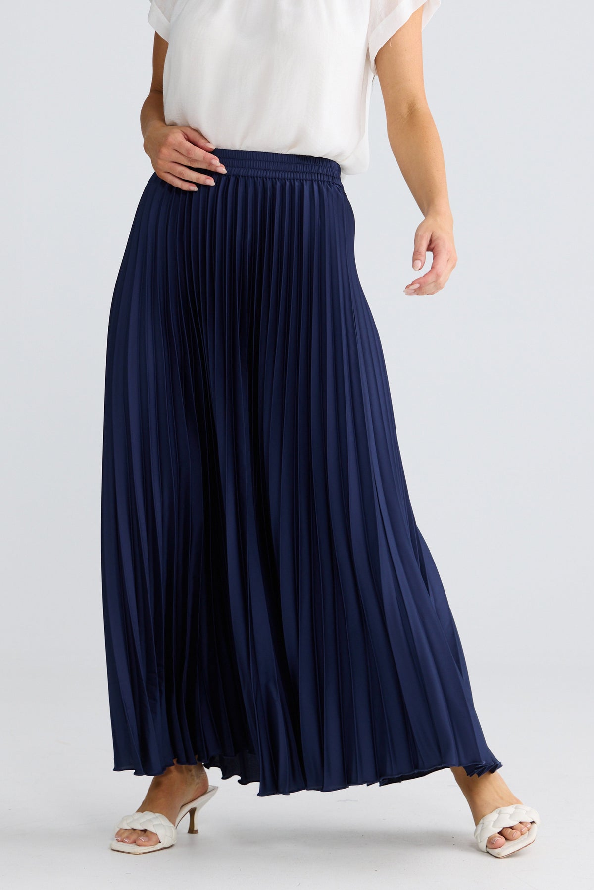 A navy blue pleated maxi skirt worn by a model, styled with a white sleeveless top and white heeled sandals against a light gray studio background.