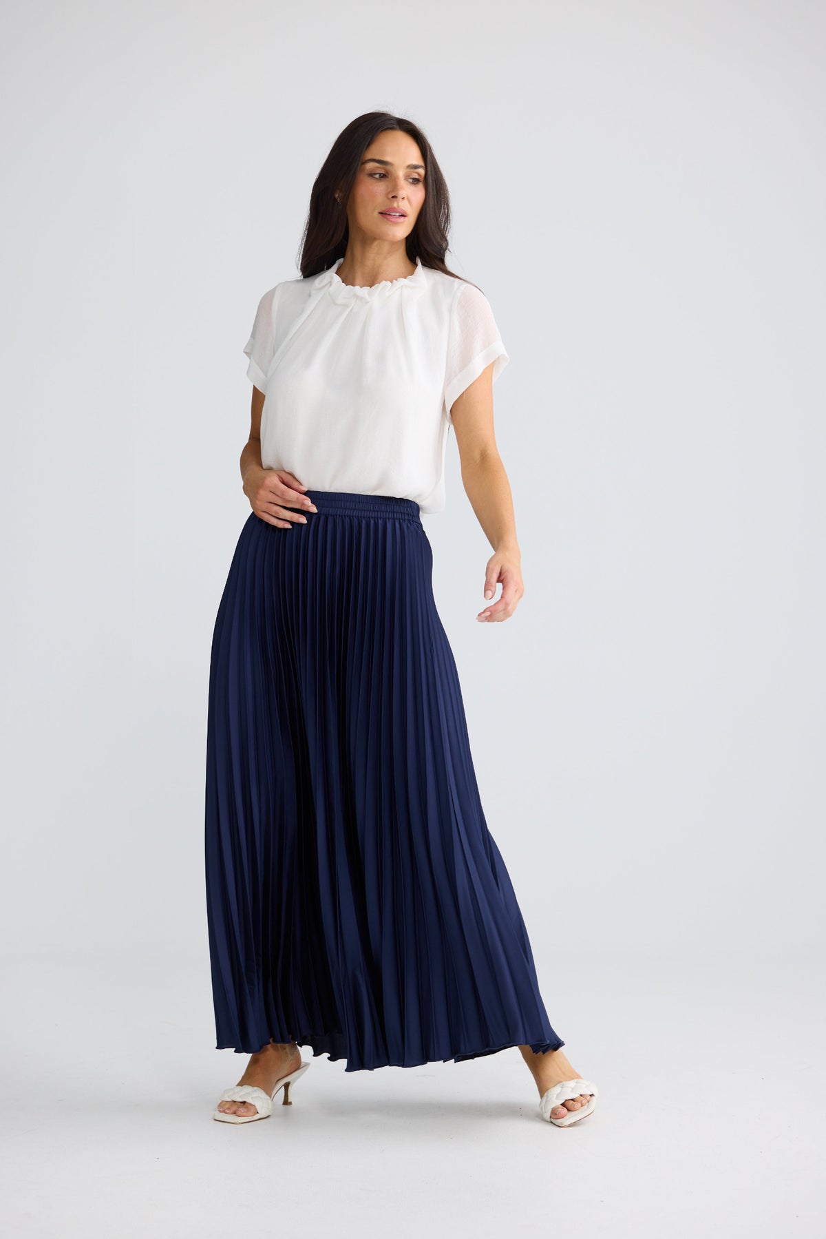 A woman modeling a white short-sleeve blouse and a navy blue pleated maxi skirt against a light gray studio background.