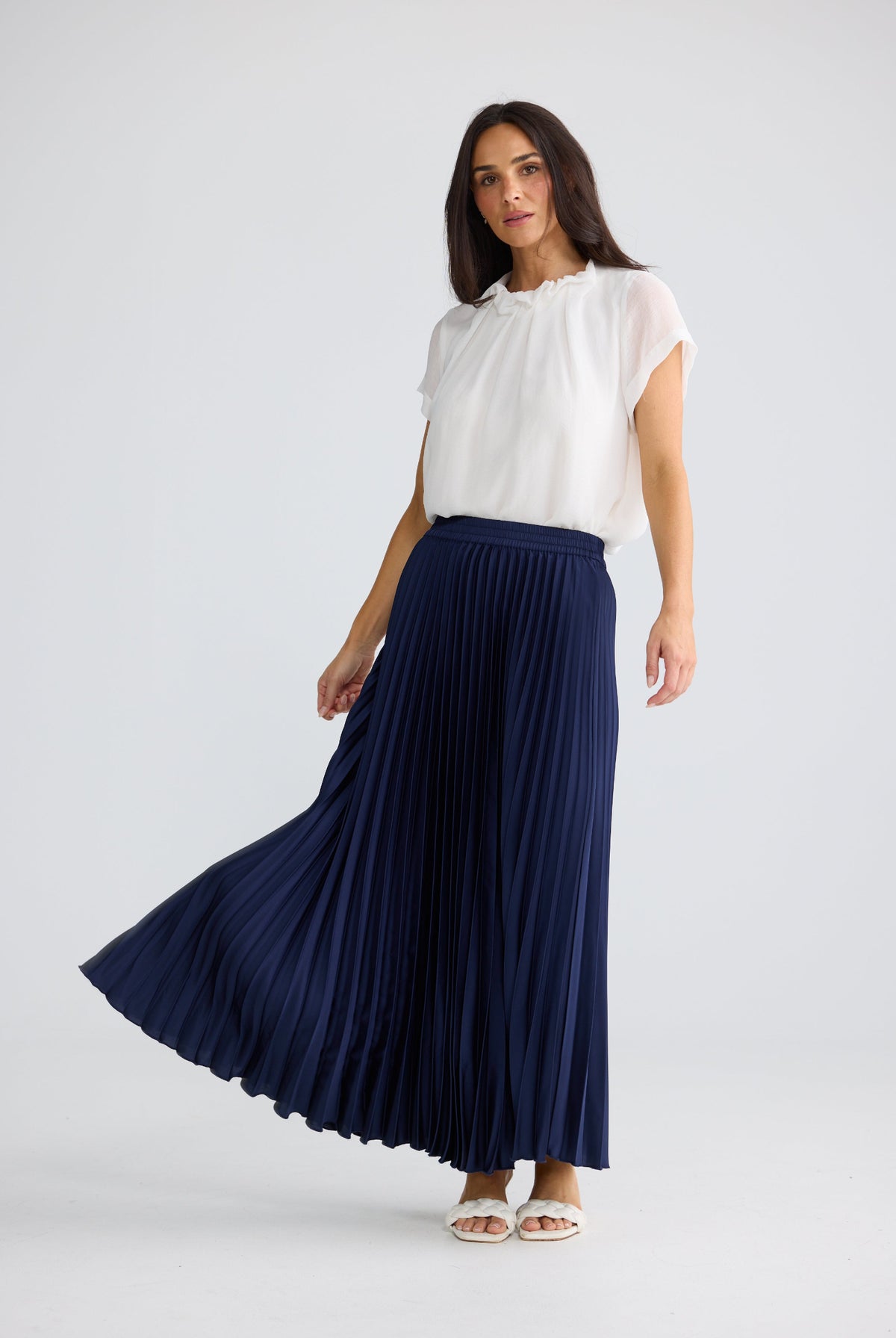 A woman modeling a navy blue pleated maxi skirt paired with a white short-sleeve blouse, standing against a light gray studio background.