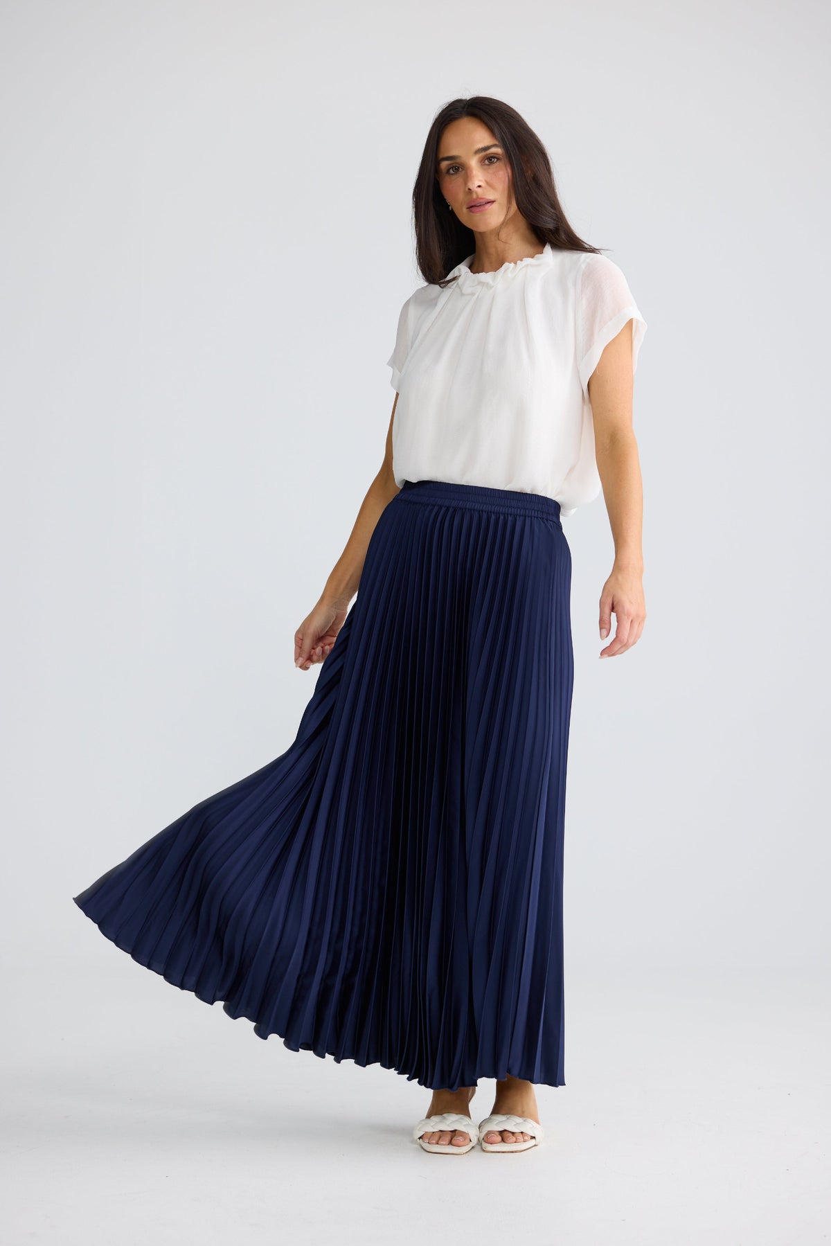 A woman modeling a navy blue pleated maxi skirt paired with a white short-sleeve blouse, standing against a light gray studio background.