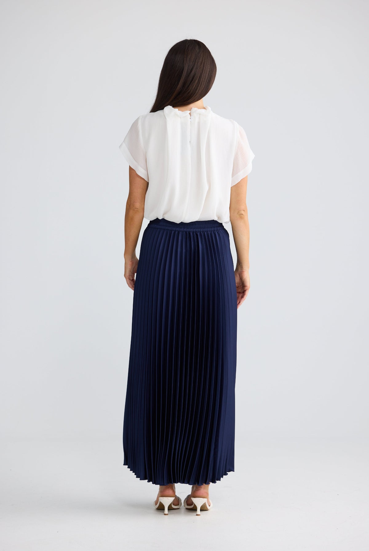 A woman shown from the back wearing a white short-sleeve blouse and a navy blue pleated maxi skirt, standing against a light gray studio background.