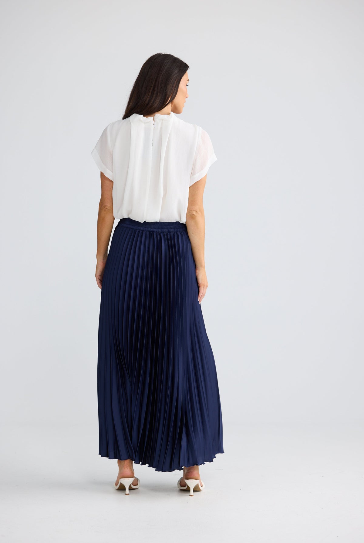A woman shown from the back wearing a white short-sleeve blouse and a navy blue pleated maxi skirt, standing against a light gray studio background.