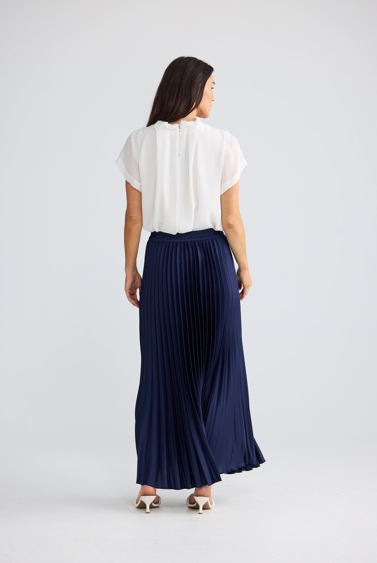 A woman shown from the back wearing a white short-sleeve blouse and a navy blue pleated maxi skirt, standing against a light gray studio background.