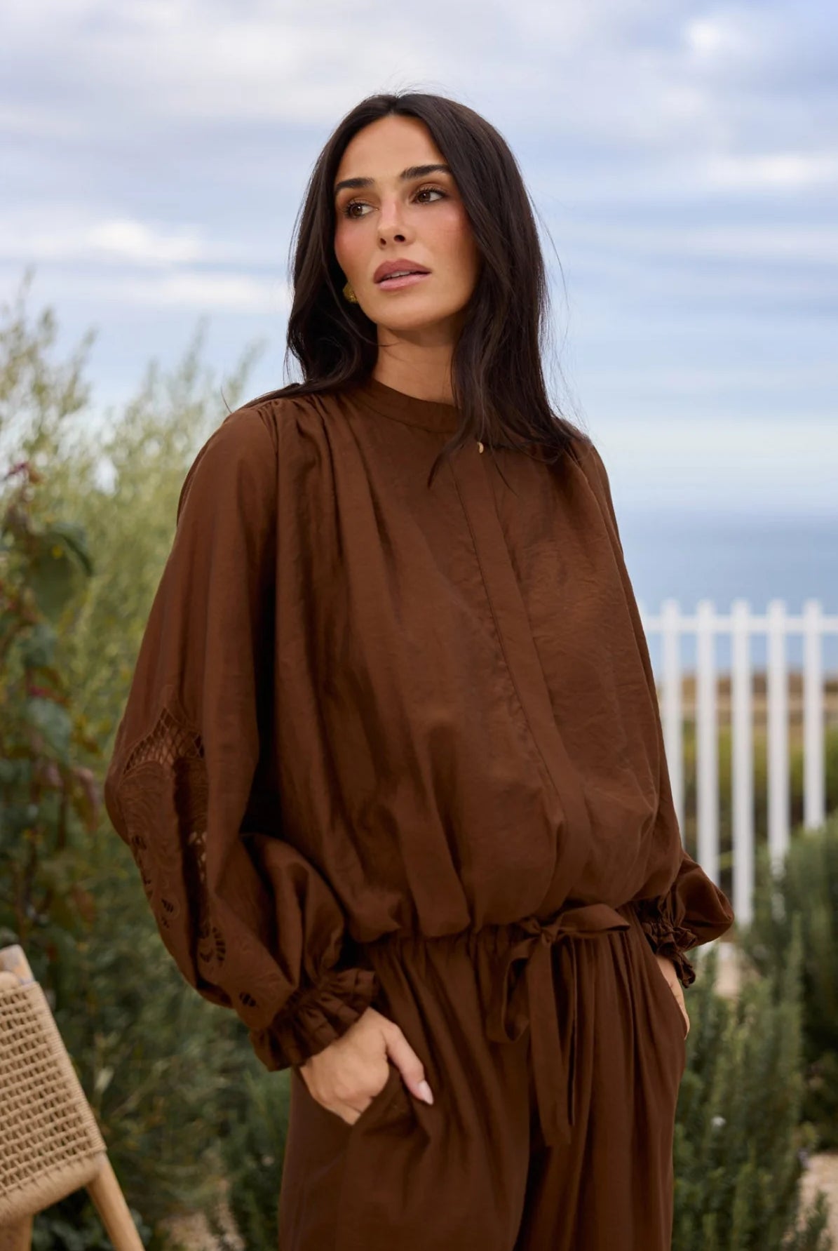 A woman wearing a brown long-sleeve top and pant with a tie waist, standing outdoors in front of a white fence and greenery.
