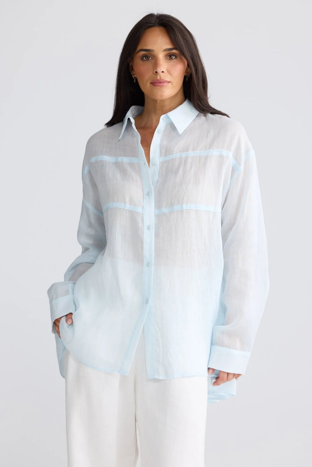 A woman modeling a light blue and white semi-sheer button-up shirt with light blue stripe details, paired with white pants, against a light gray studio background.