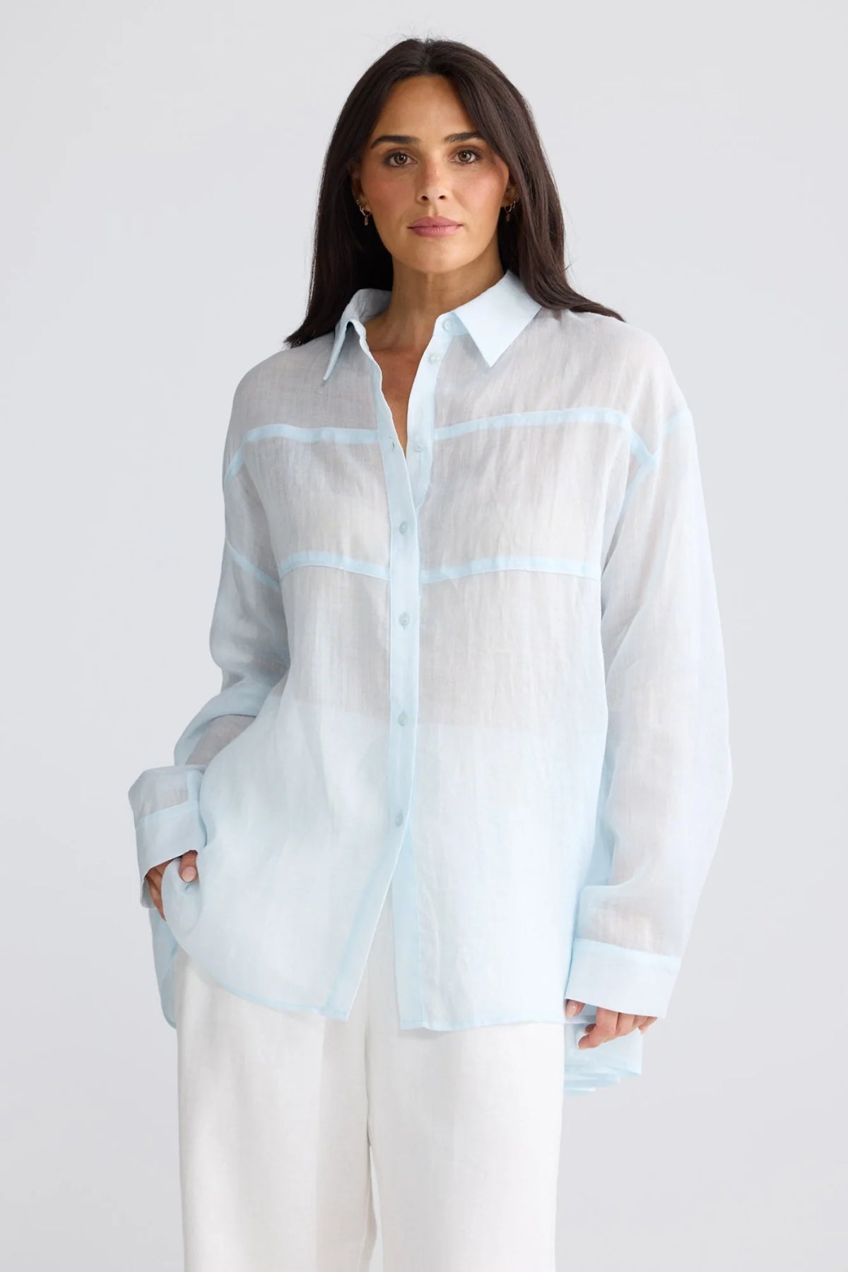 A woman modeling a light blue and white semi-sheer button-up shirt with light blue stripe details, paired with white pants, against a light gray studio background.