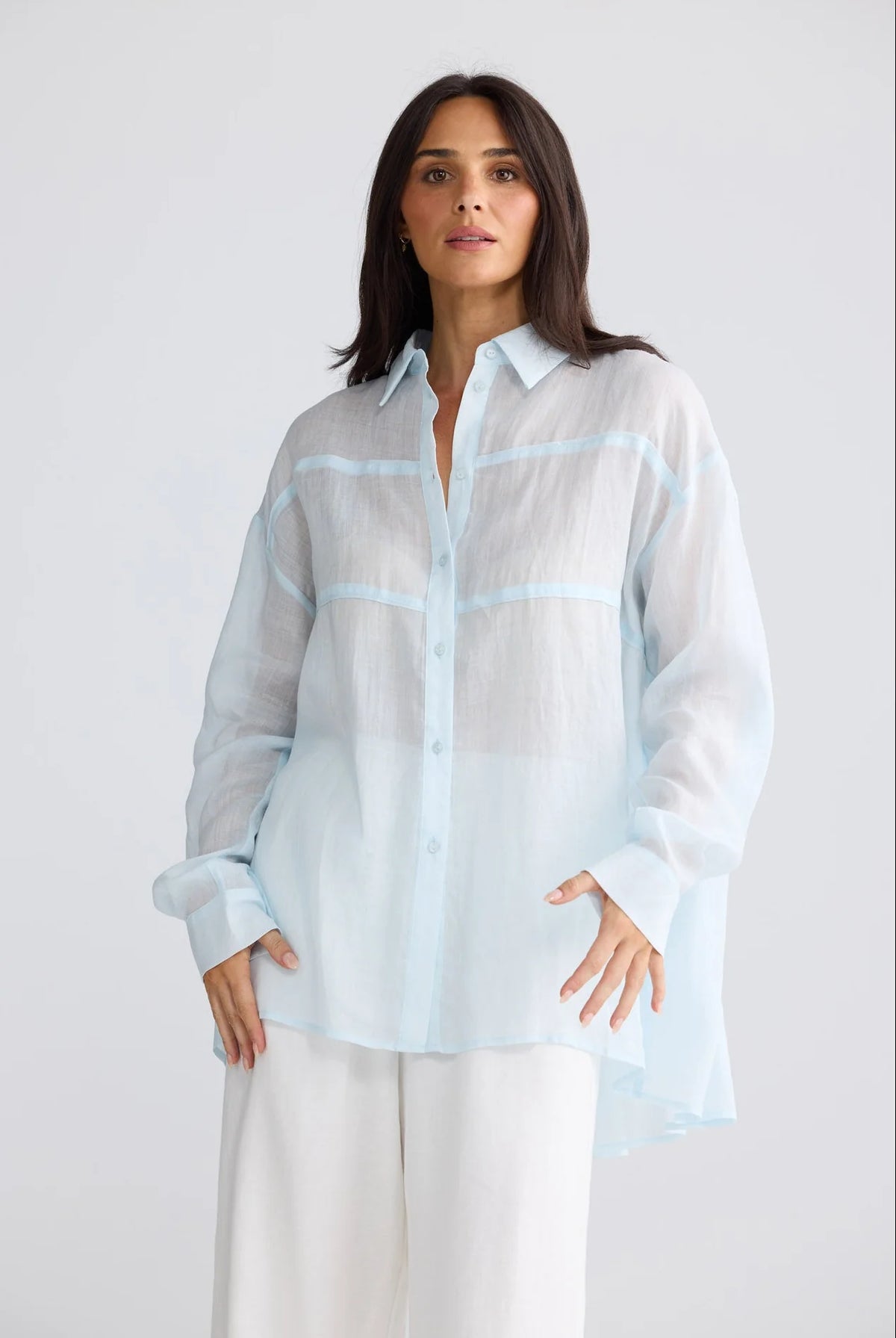 A woman modeling a light blue semi-sheer long-sleeve button-up shirt with white wide-leg pants, photographed against a clean light gray studio background.