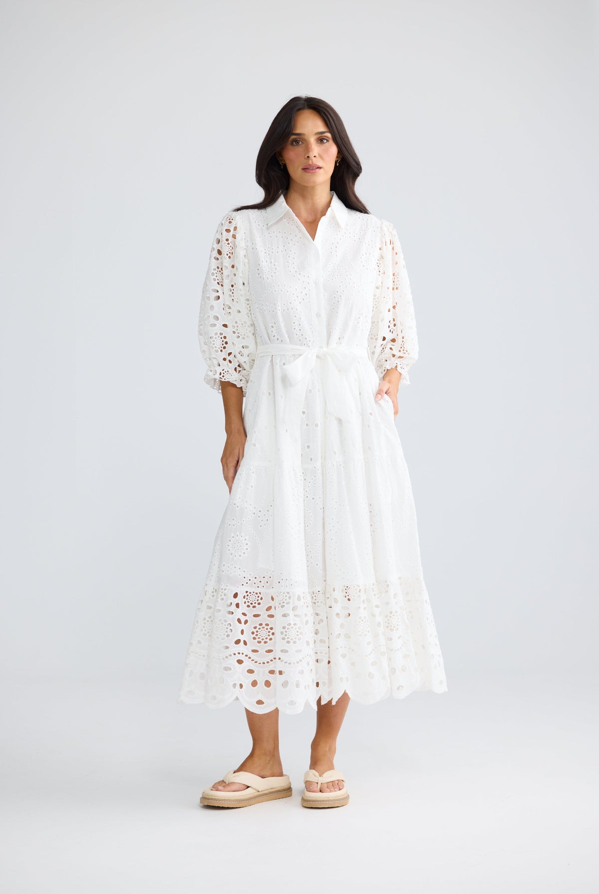 A woman modeling a white eyelet midi dress with three-quarter sleeves, a tie waist, and side pockets, paired with beige espadrille sandals against a light gray studio background.