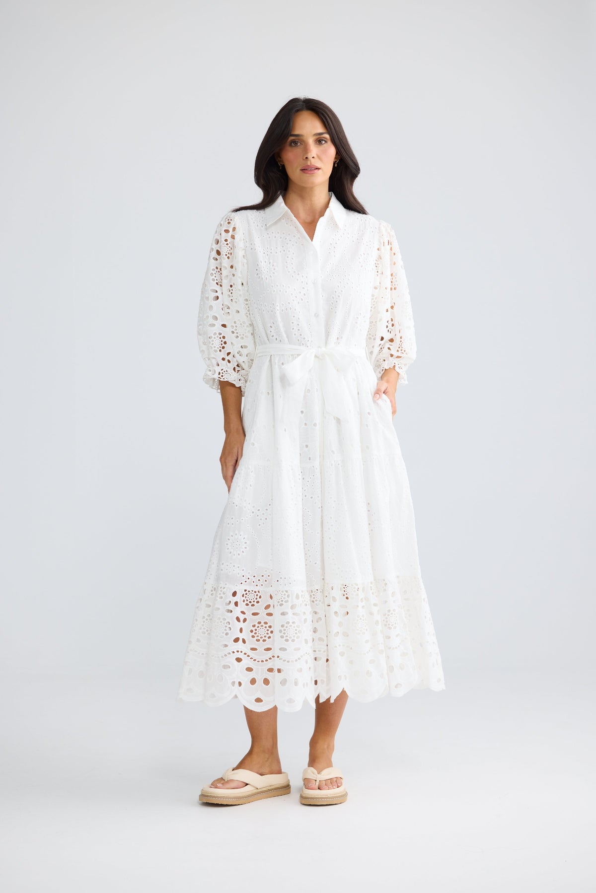A woman modeling a white eyelet midi dress with three-quarter sleeves, a tie waist, and side pockets, paired with beige espadrille sandals against a light gray studio background.