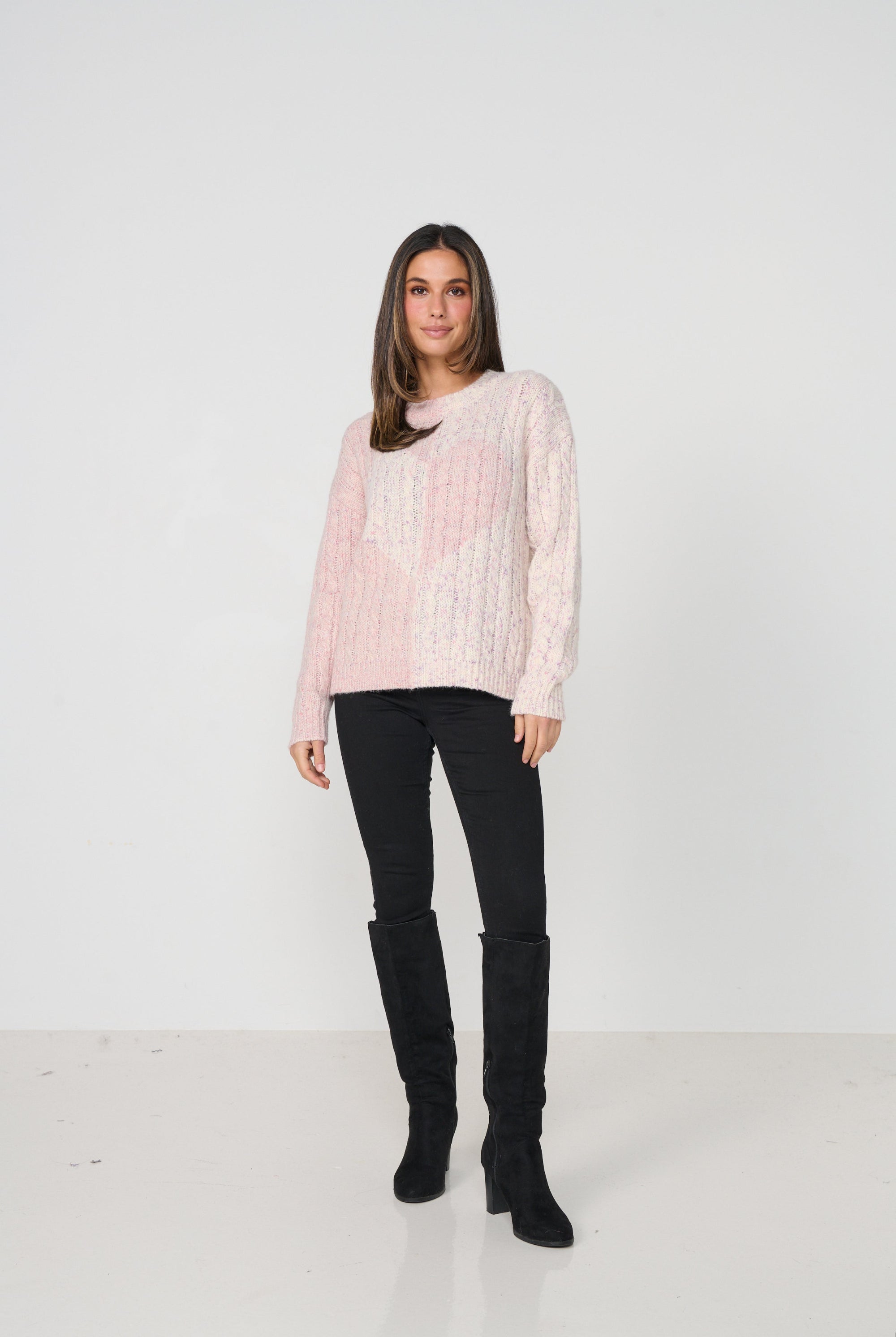 Woman wearing a light pink cable knit sweater and black pants on a white background