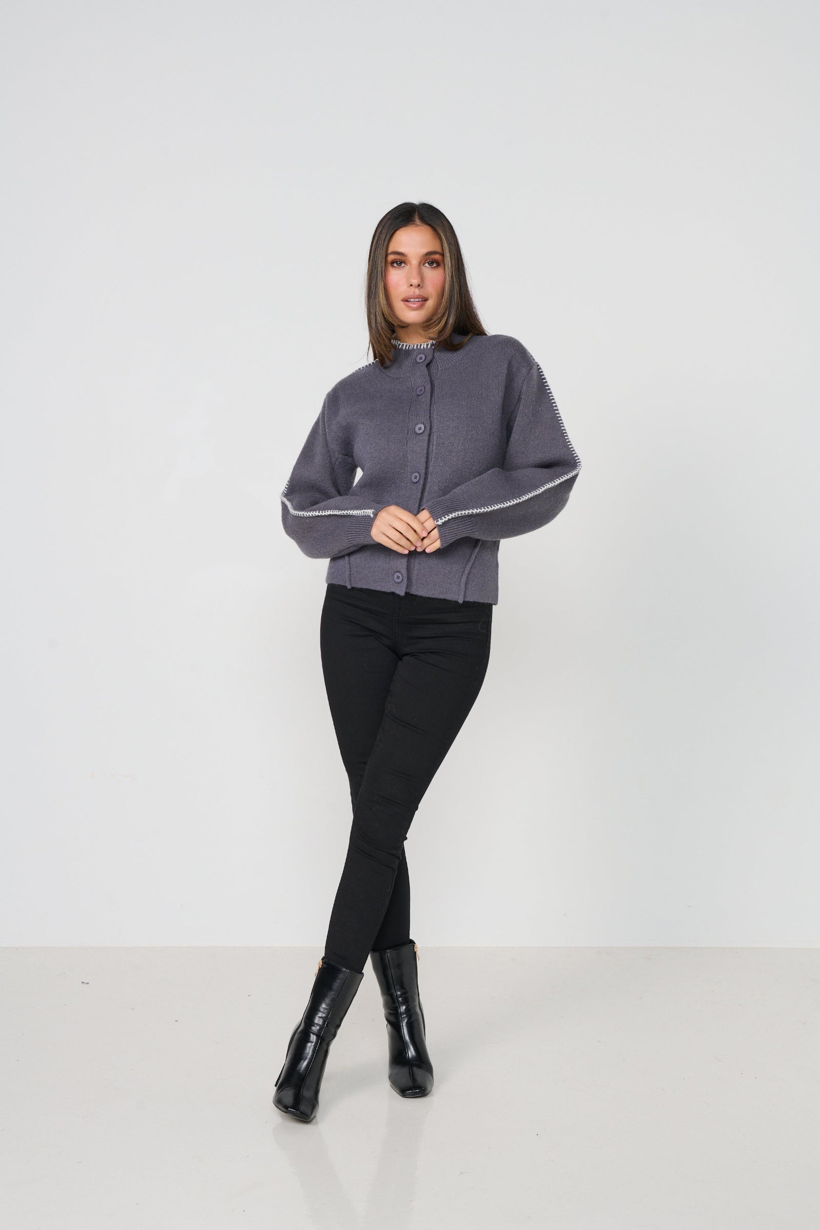 Woman wearing a gray cardigan with white trim, black pants, and black boots on a white background