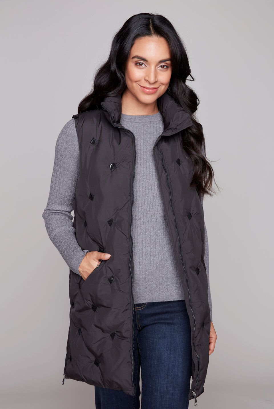 Woman wearing a black longline puffer vest over a gray sweater on a plain background