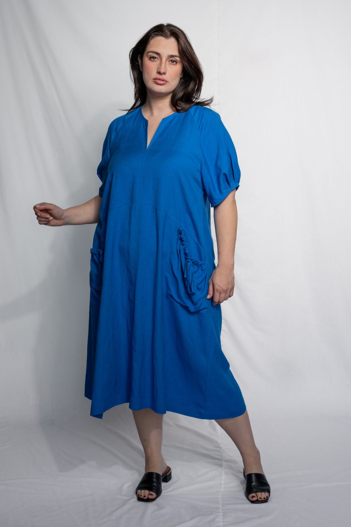 A woman modeling a bright blue midi dress with short puff sleeves, a split neckline, and large side pockets, standing against a white studio backdrop.