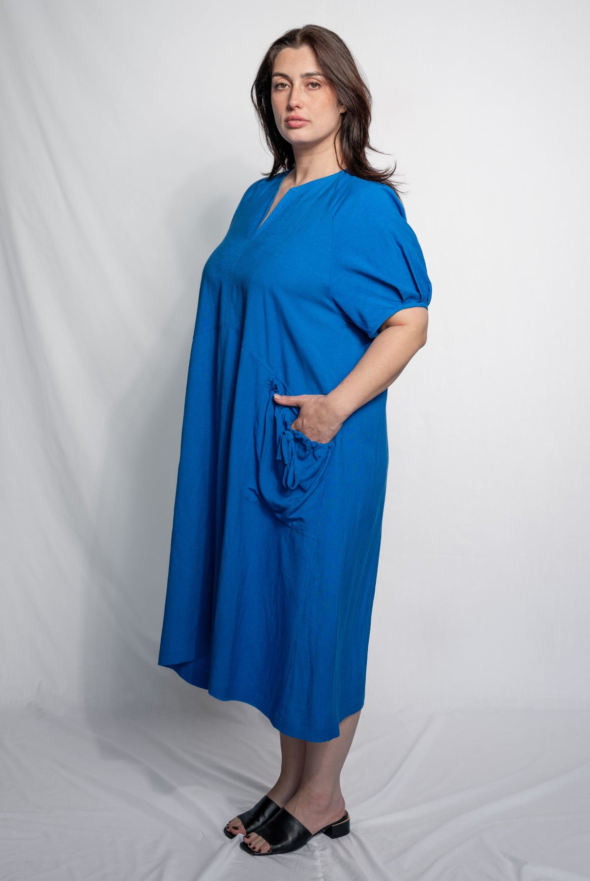 A woman modeling a bright blue midi dress with short sleeves, a split neckline, and a side pocket, standing against a white studio backdrop.