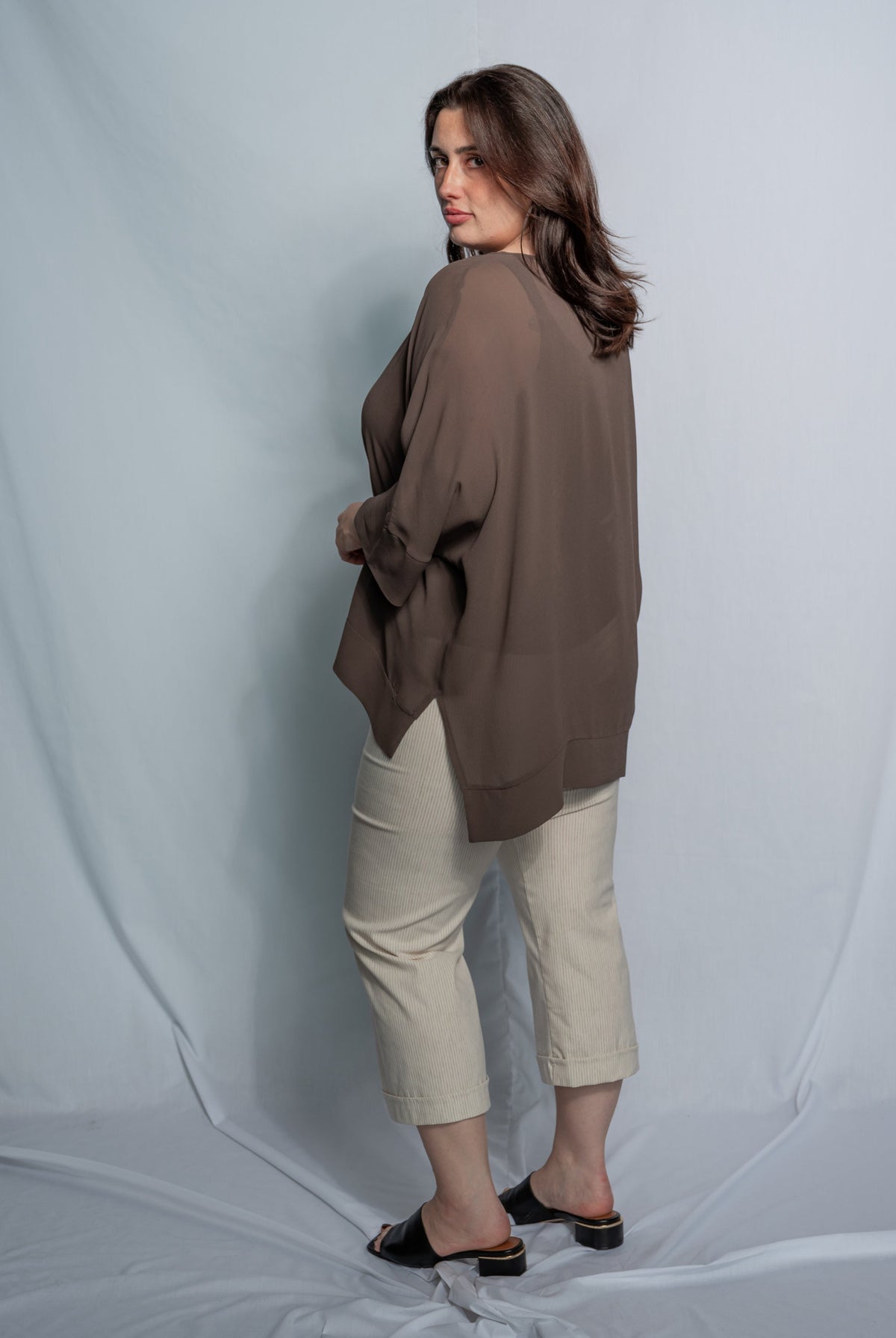 A woman modeling a loose, oversized taupe-brown top paired with beige cropped pants and black slide sandals, standing against a light gray studio backdrop.