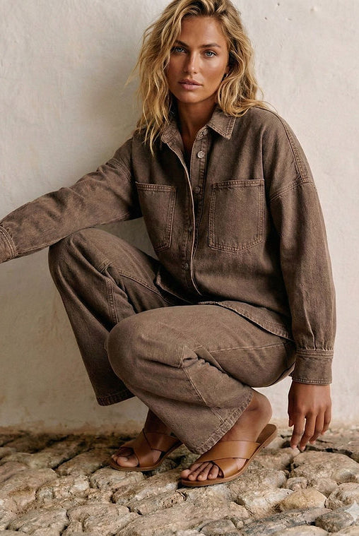 A woman crouches against a textured white wall on a stone surface, wearing a matching brown corduroy button-up shirt and wide-leg pants set with tan leather slide sandals.