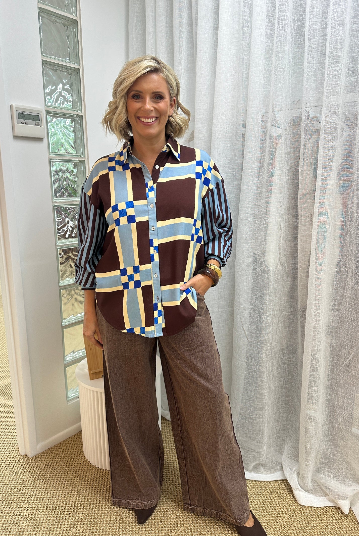 A woman posing indoors wearing a brown and navy striped button-up shirt with a blue, yellow, and brown geometric patterned vest, paired with brown wide-leg pants.